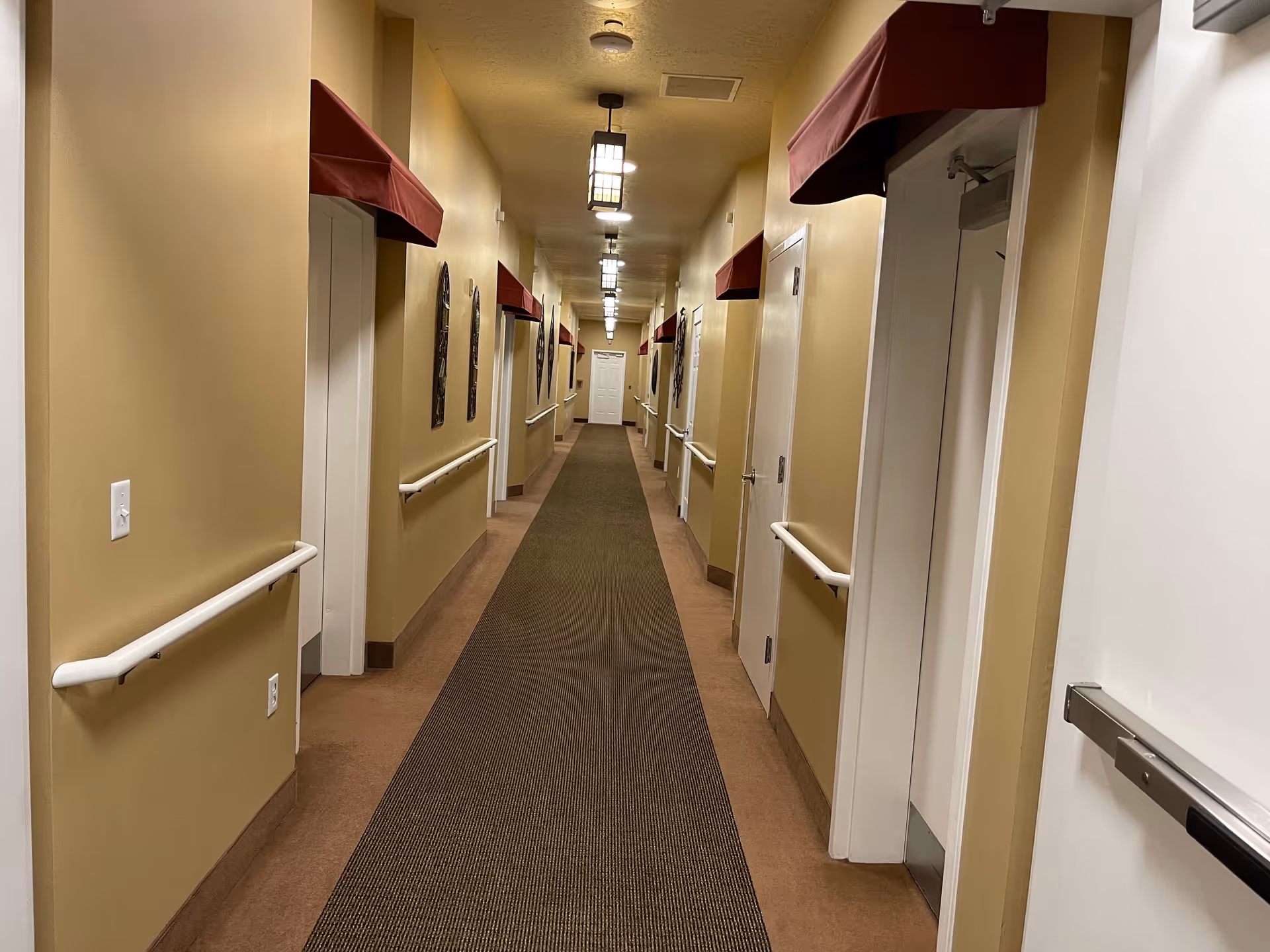 A long, narrow hallway in a senior living facility with beige walls and a brown carpet runner. The hallway has white handrails on both sides and several doors with red awnings above them. Ceiling lights are evenly spaced along the corridor, and decorative wall hangings are visible on the left side.