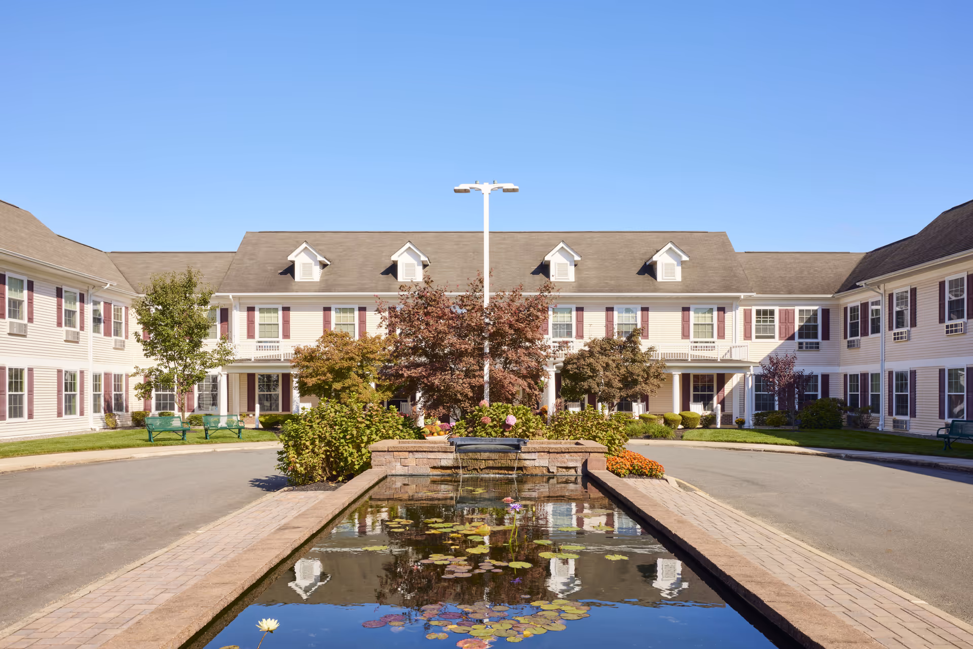 Exterior view of Brandywine Toms River by Monarch senior living facility showing a U-shaped building with beige siding and maroon shutters. In front is a rectangular pond with water lilies and a small waterfall, surrounded by landscaped bushes and trees under a clear blue sky.