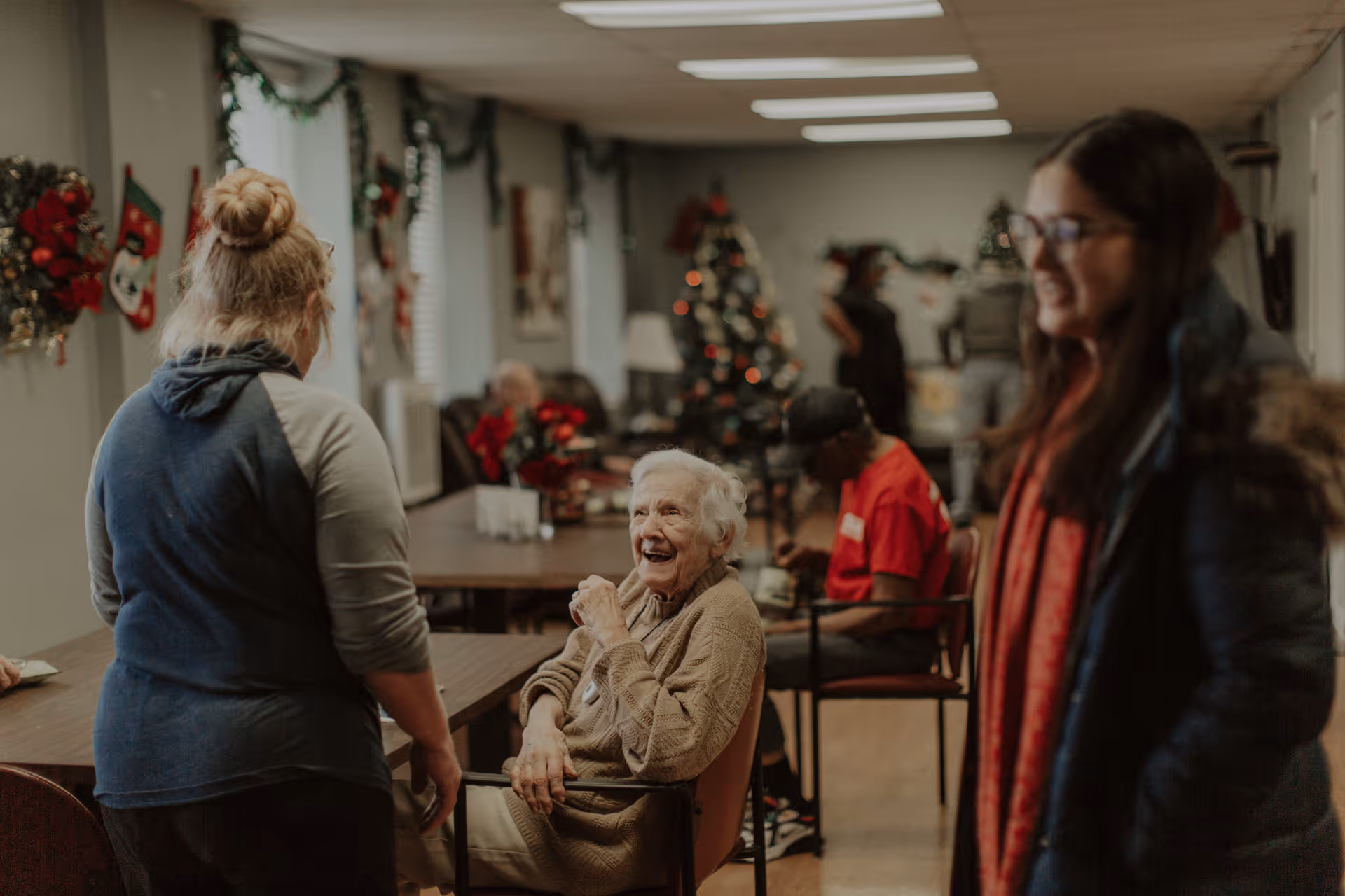 An elderly woman laughs while residents and visitors socialize in a decorated common room with a Christmas tree.