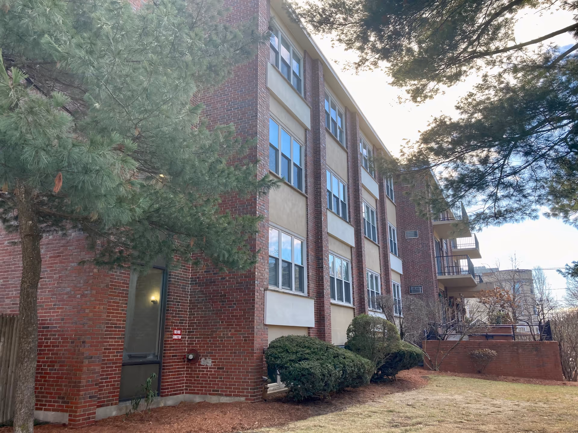 Exterior view of a multi-story brick building with several windows and balconies, surrounded by trees and bushes under a partly cloudy sky.