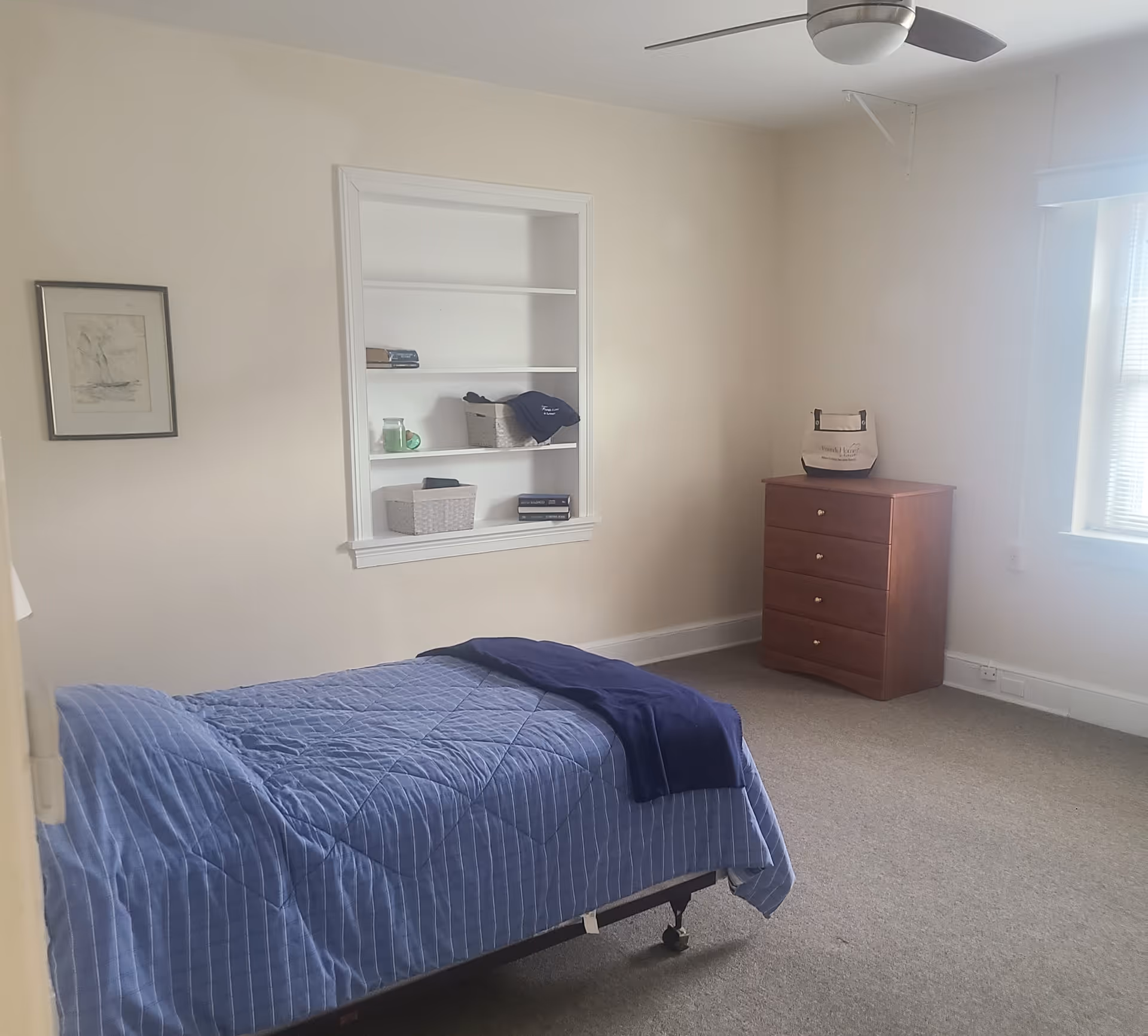 Sunny single-occupancy bedroom with a blue-covered single bed, built-in wall shelves, a wooden dresser, and a window.