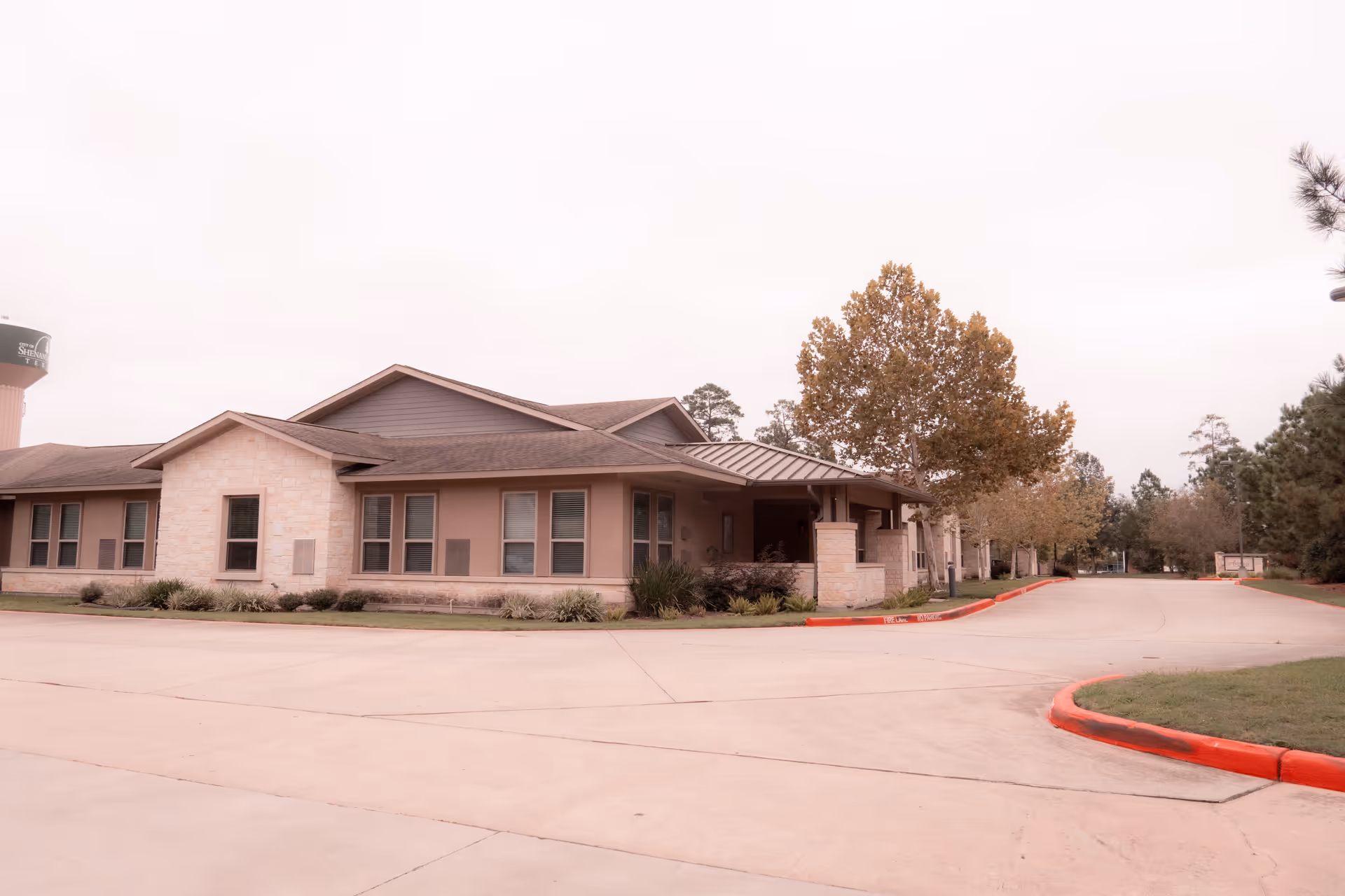 Single-story senior living building with a covered entrance, driveway, and trees under an overcast sky.