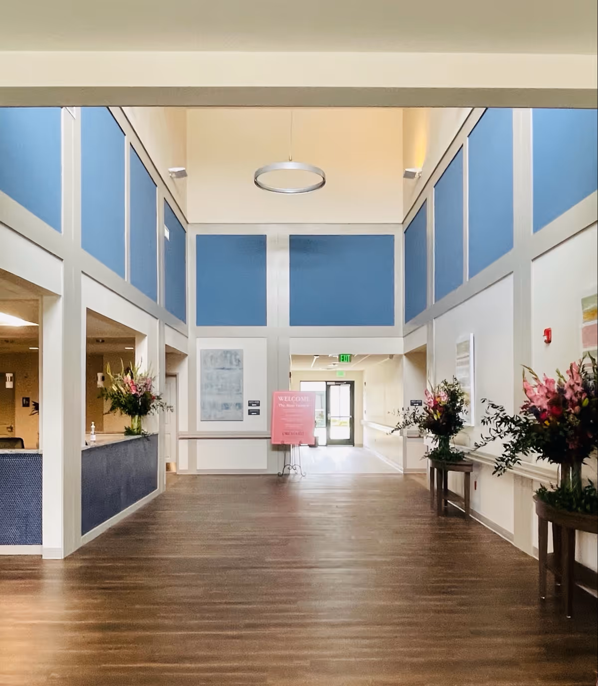 Interior view of a senior living facility lobby with high ceilings and blue and white paneled walls. There is a modern circular light fixture hanging from the ceiling. The floor is wooden, and there are flower arrangements on tables along the right wall and on the reception desk to the left. A pink welcome sign is visible in the distance near the entrance door.