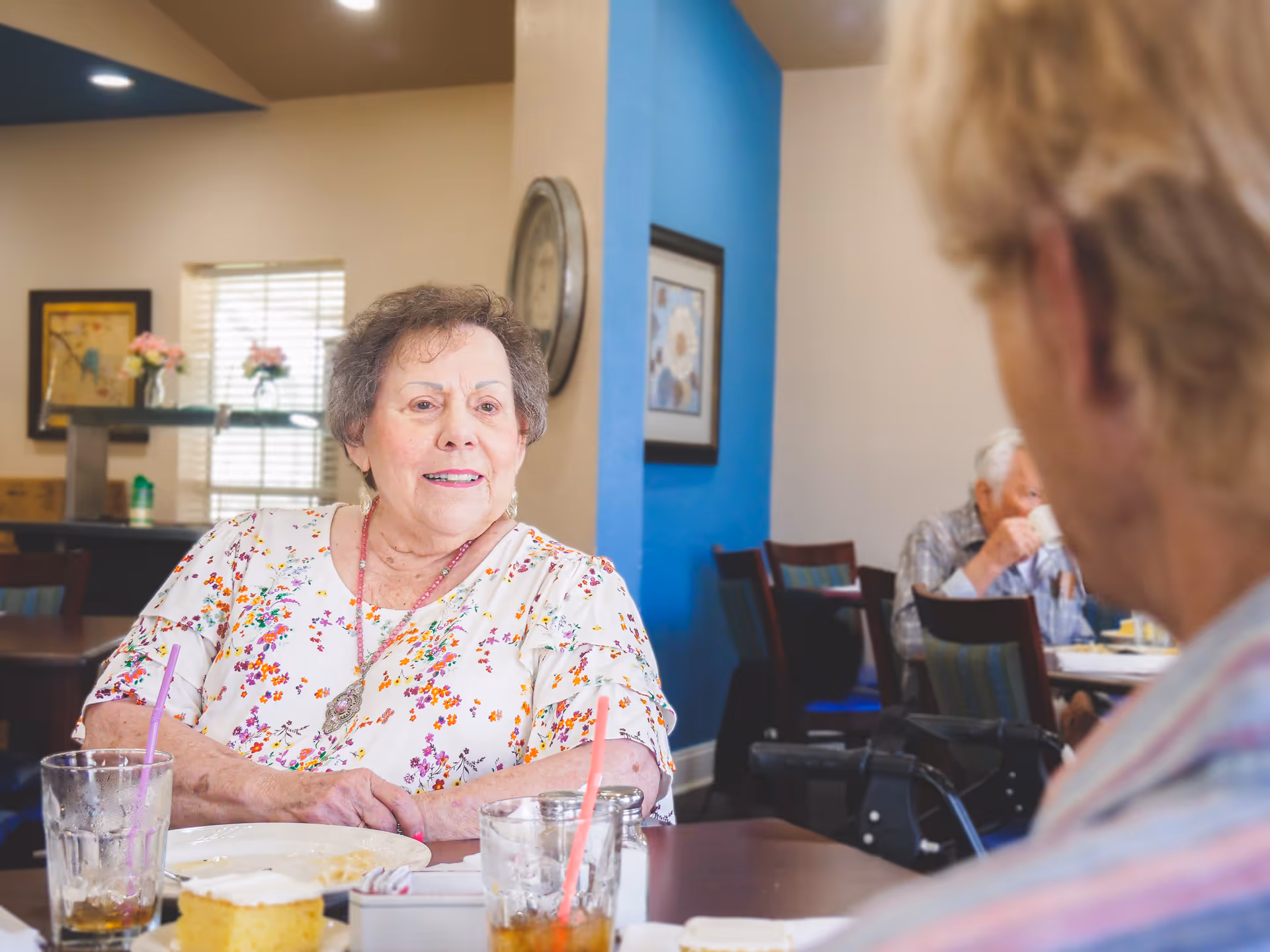 An elderly woman with short curly hair wearing a floral blouse is sitting at a dining table in a senior living facility, talking to another person whose back is to the camera. There are glasses with straws, a plate with food, and a slice of cake on the table. In the background, another elderly person is drinking from a cup in a room with blue and beige walls and framed artwork.