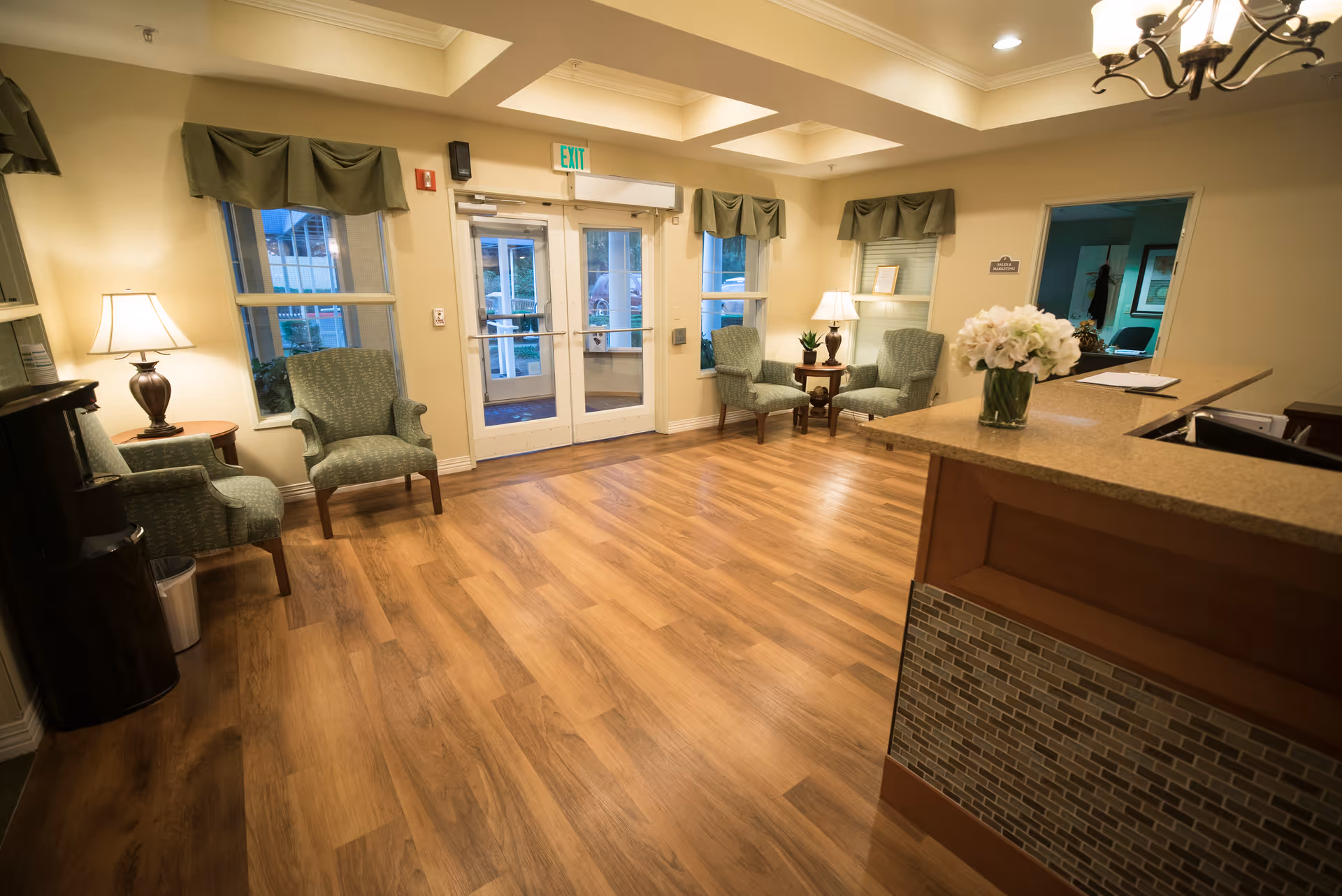 Bright senior living reception lobby with wood floors, seating near double glass doors and a front desk with a vase of flowers.