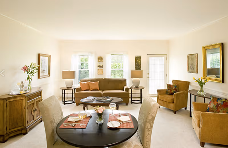 A bright and cozy living room with a round dining table set for two in the foreground. The room features a beige sofa with two orange pillows, two armchairs with colorful cushions, two side tables with lamps, a wooden cabinet with flowers and glassware, and a large mirror on the wall. There are three windows and a glass door letting in natural light.