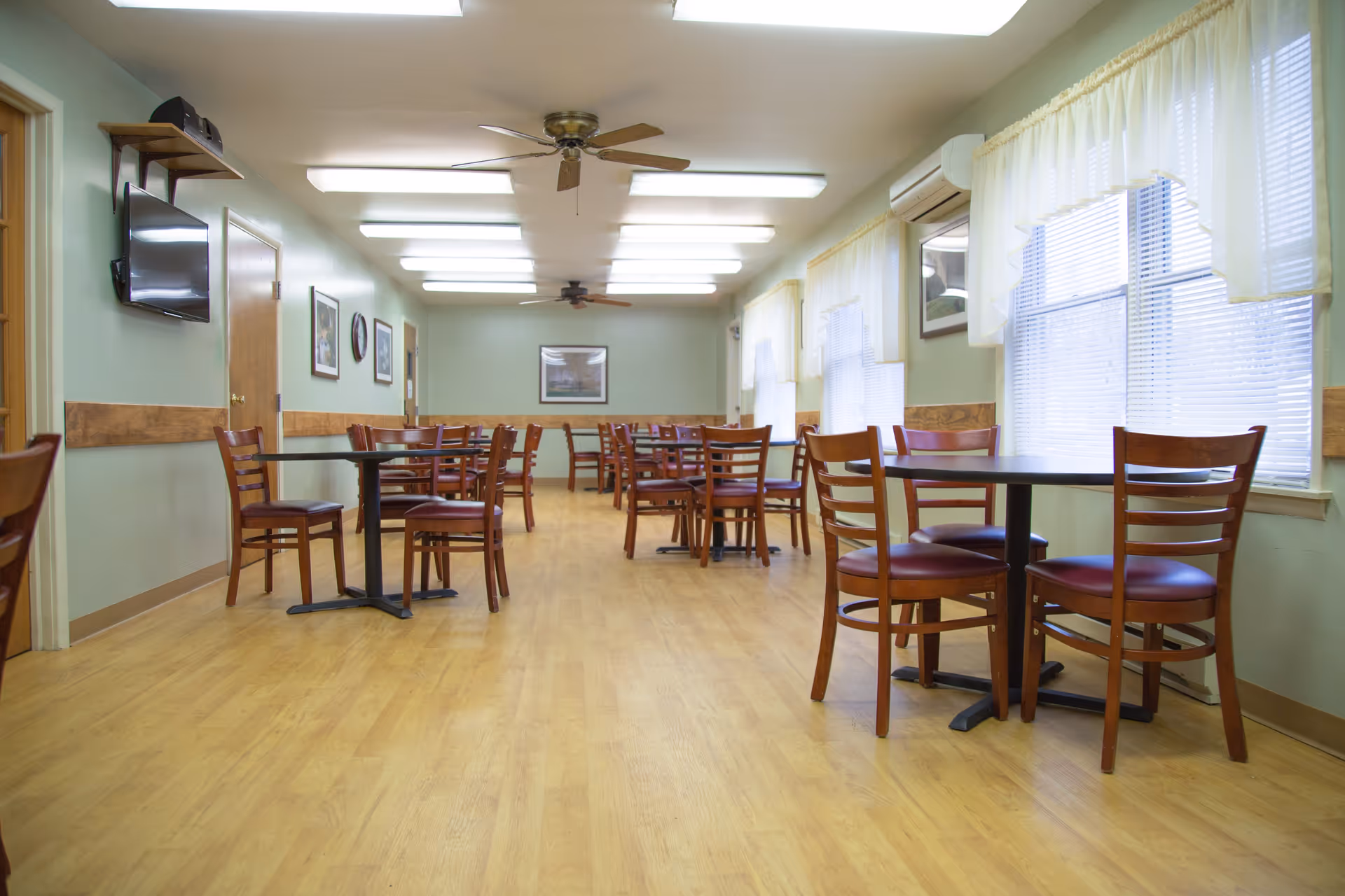 A dining room with multiple wooden tables and chairs arranged neatly on a light wood floor. The room has light green walls with wooden trim, several windows with white curtains, ceiling fans, and fluorescent lighting. A wall-mounted TV and framed pictures are visible on the walls.