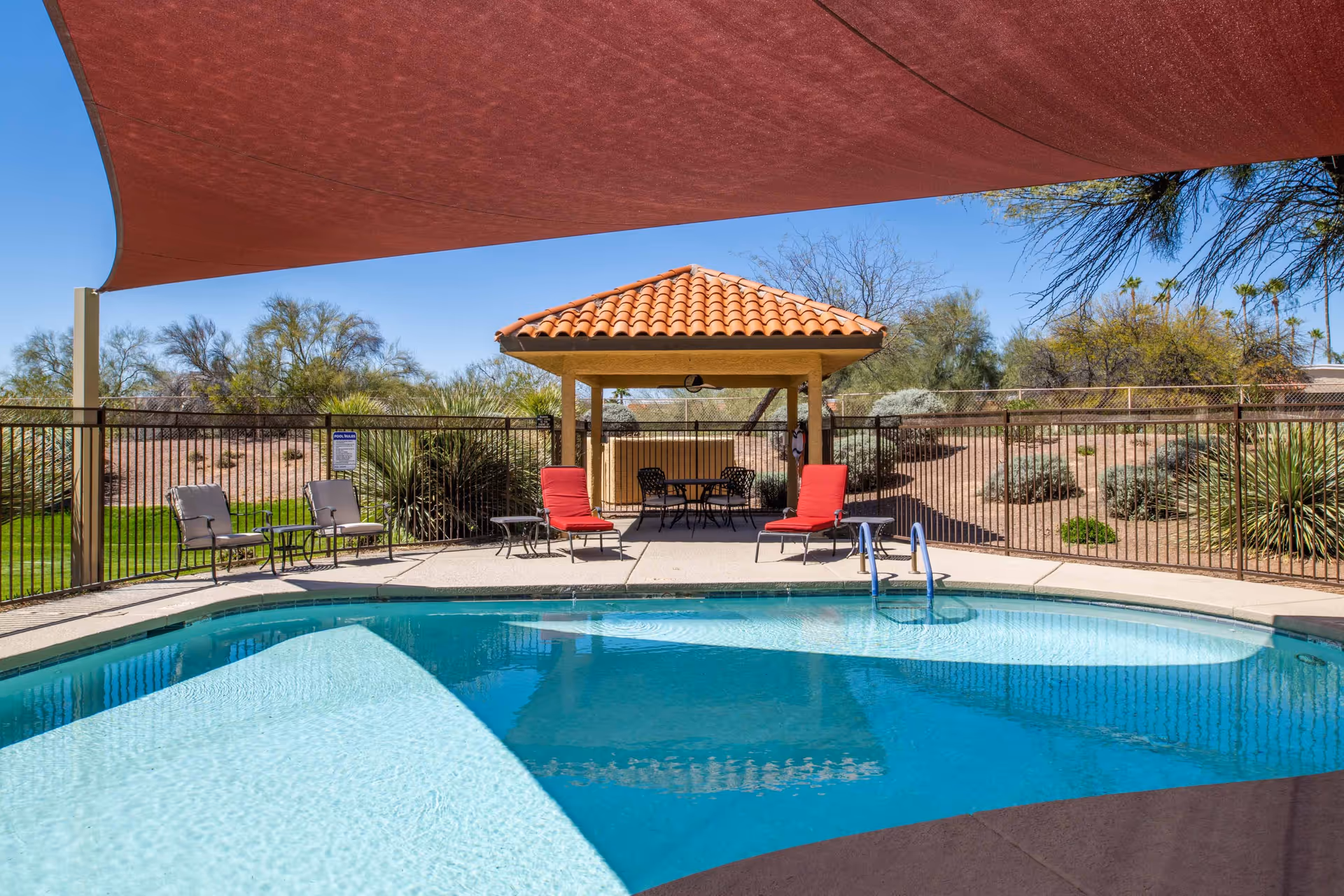 Outdoor swimming pool with clear blue water, surrounded by a concrete deck with lounge chairs and small tables. A shaded canopy covers part of the pool area, and a small gazebo with a tiled roof and seating is visible in the background. Desert landscaping with bushes and trees surrounds the fenced pool area under a clear blue sky.