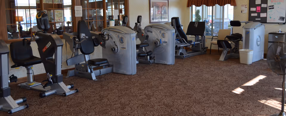 A fitness room with several exercise machines including stationary bikes and strength training equipment, carpeted floor, large windows with valance curtains, and a wall mirror reflecting the equipment.