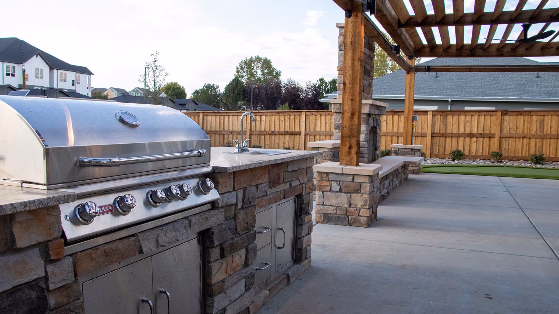 Outdoor covered patio with a built-in stainless steel grill, stone countertop and sink under a wooden pergola.