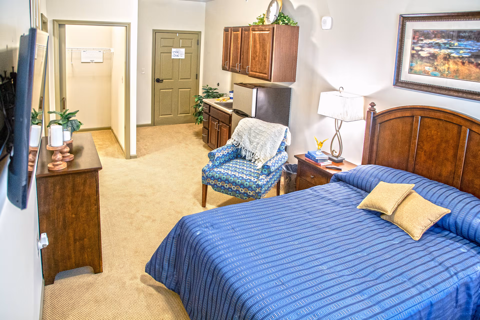 Furnished bedroom with a blue bedspread, patterned armchair, nightstand, wall-mounted TV and small kitchenette in a senior living unit.