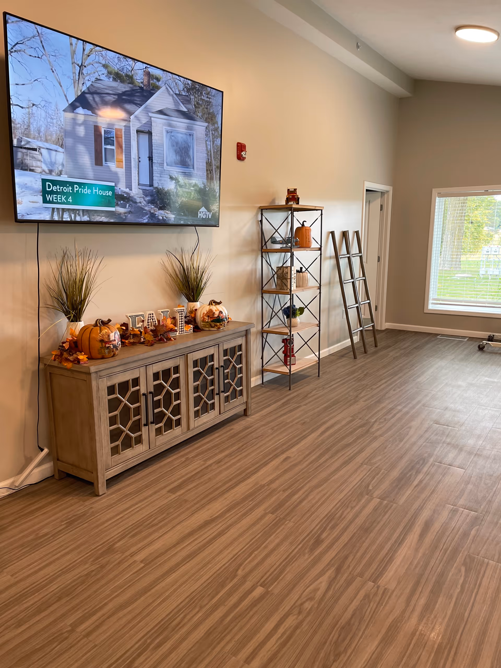 A spacious room with wood-patterned flooring and beige walls. A large flat-screen TV is mounted on the wall displaying a show titled 'Detroit Pride House WEEK 4' on HGTV. Below the TV is a wooden cabinet decorated with fall-themed items including pumpkins, plants, and a sign that reads 'FALL'. To the right of the cabinet is a metal and wood shelving unit with decorative items and a folded ladder leaning against the wall. A large window with blinds lets in natural light from outside.