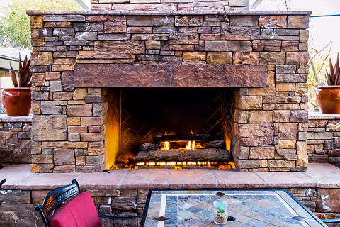Outdoor stone fireplace with a fire burning inside, flanked by two potted plants on stone ledges. In front of the fireplace is a table with a mosaic tile top and a chair with a red cushion.