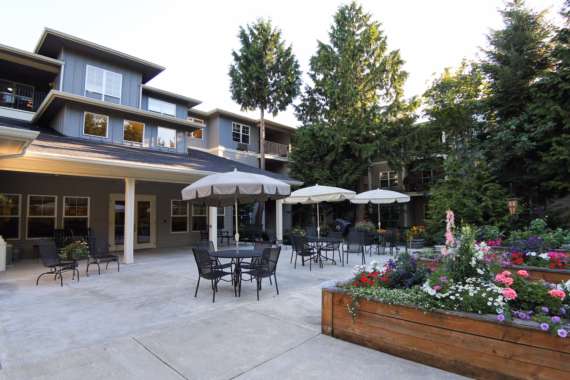 Courtyard patio with tables, umbrellas, metal chairs, wooden planter boxes filled with colorful flowers, and a multi-story residential building in the background.