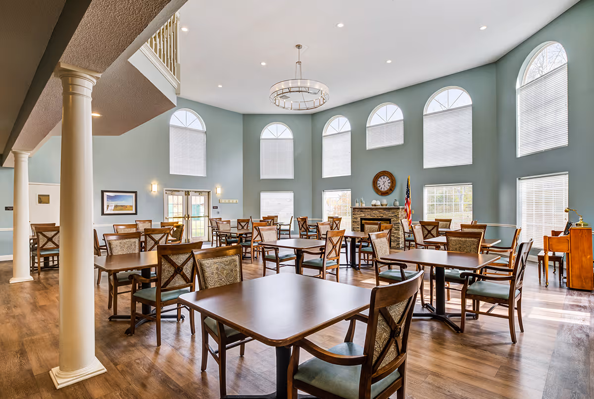 A spacious dining room with multiple wooden tables and chairs arranged neatly on a wooden floor. The room features high ceilings with large arched windows covered with blinds, a modern chandelier, and a stone fireplace with a clock above it. An American flag stands near the fireplace, and there is a small wooden piano in the corner.