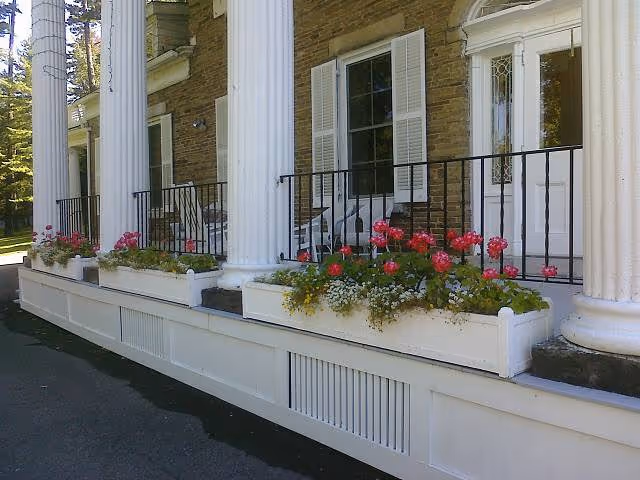 Front exterior view of a building with large white columns, white window shutters, a white door with decorative glass, and flower boxes filled with pink and white flowers along the porch railing.