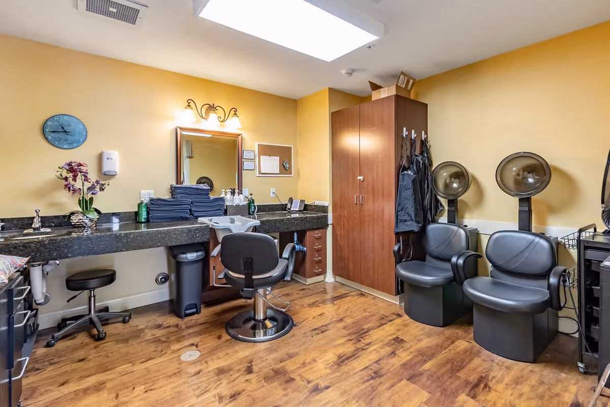Interior view of a salon room with yellow walls and wood flooring. The room features a long black countertop with a sink, a mirror, and a stack of folded towels. There is a salon chair in front of the sink and two black hair dryer chairs with hooded dryers on the right side. A wooden cabinet and several hanging aprons are also visible.