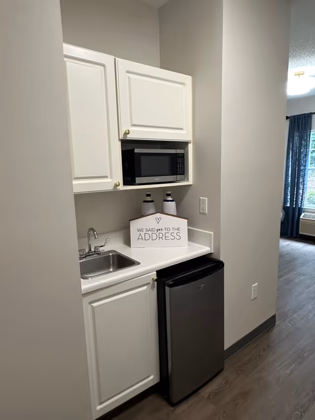 A small kitchenette area with white cabinets, a stainless steel sink, a microwave, and a mini refrigerator. On the countertop, there is a decorative sign that reads 'We said yes to the address' along with two decorative bottles. The kitchenette is adjacent to a room with a window and blue curtains.