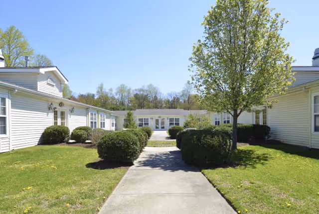 A paved walkway leading through a landscaped courtyard with trimmed bushes and a tree, surrounded by single-story white buildings under a clear blue sky.