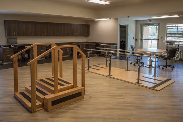 A rehabilitation therapy room with wooden stairs for physical therapy and parallel bars for walking exercises. The room has wooden flooring, office chairs, desks with computers, and an exit door with windows letting in natural light.