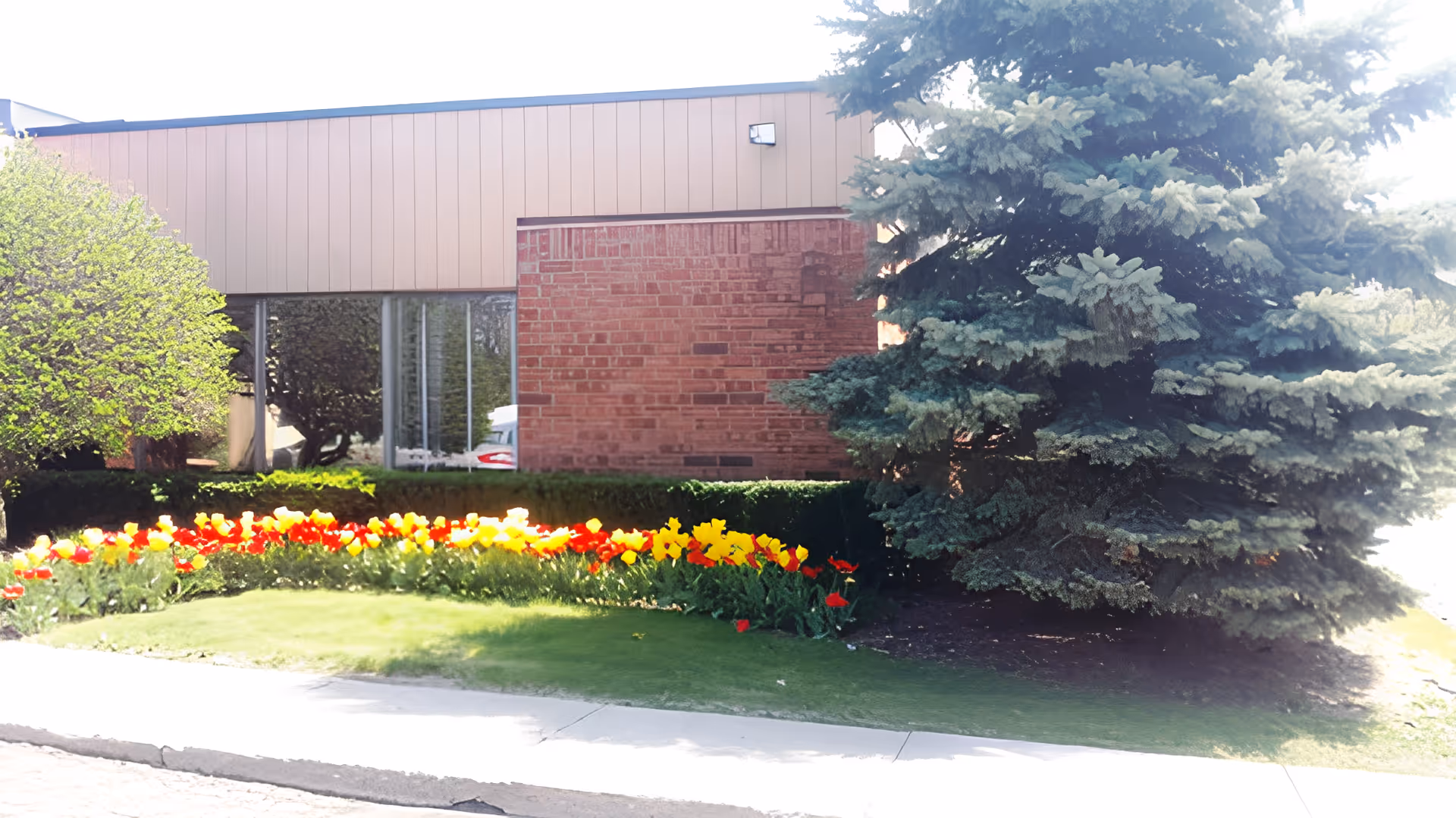 Exterior view of a building with a brick and tan panel facade, surrounded by a landscaped garden featuring colorful red and yellow flowers, green bushes, and a large evergreen tree.