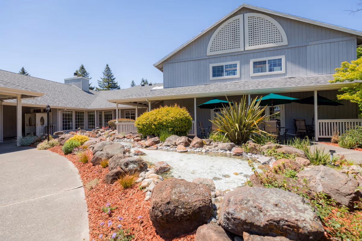 Exterior view of Cogir of Rohnert Park facility showing a landscaped garden with rocks, plants, and a small water feature in front of a gray building with a covered patio area furnished with tables and green umbrellas under a clear blue sky.
