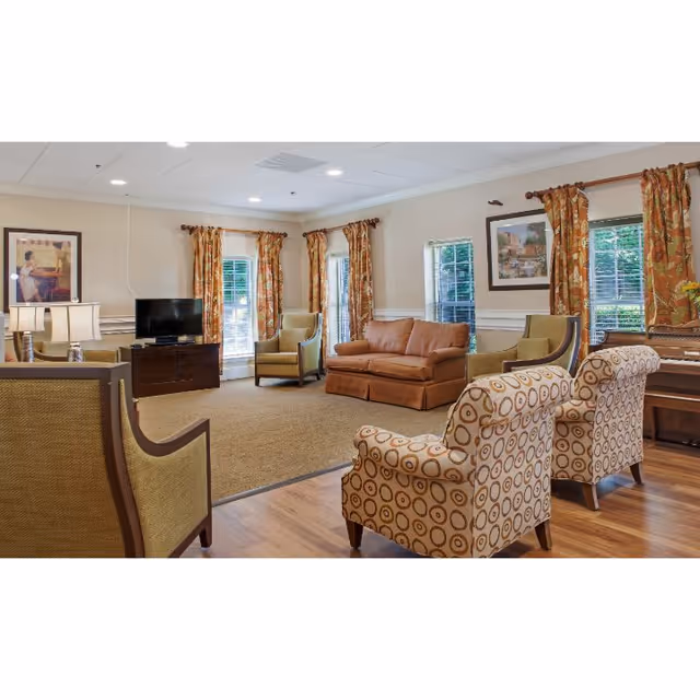 A cozy living room area with multiple armchairs and a sofa arranged around a carpeted floor. The room has large windows with patterned curtains, a flat-screen TV on a wooden stand, framed artwork on the walls, and warm lighting from table lamps.