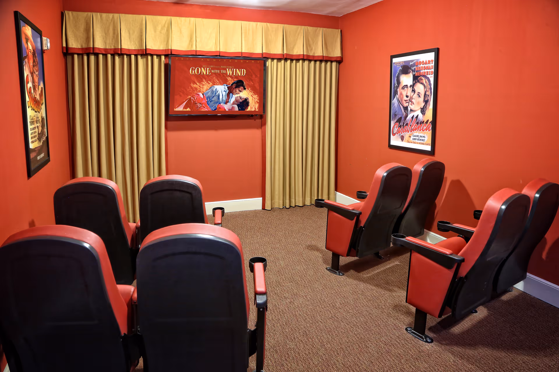Small home theater room with six red and black theater seats arranged in two rows facing a screen displaying the movie poster for Gone with the Wind. The walls are painted red and decorated with framed classic movie posters including Casablanca. Beige curtains cover the window behind the screen.