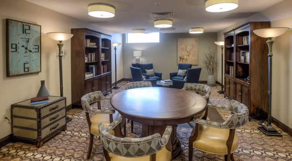 A cozy senior living common area with a round wooden table surrounded by six upholstered chairs. The room features two large wooden bookshelves filled with books, several floor lamps, a small chest with decorative items, and a seating area with four blue armchairs and a side table under a window. A painting of a deer and a tall vase with branches decorate the far wall.