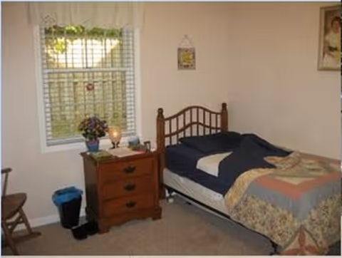 Small furnished bedroom with a single bed, wooden nightstand, window with blinds, and a chair.
