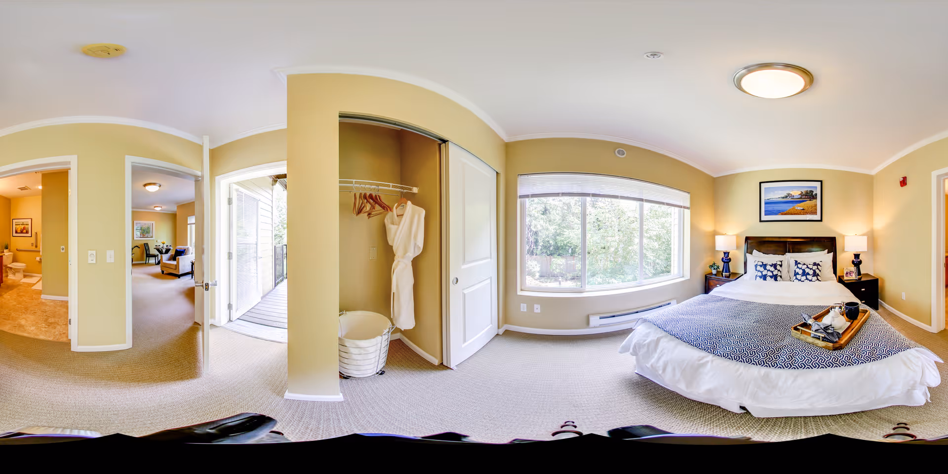 A bright and spacious bedroom in a senior living facility with a large window letting in natural light. The room features a neatly made bed with decorative pillows and a tray on top, two bedside tables with lamps, a framed picture on the wall, a closet with a bathrobe hanging inside, and an open door leading to a balcony and other rooms.