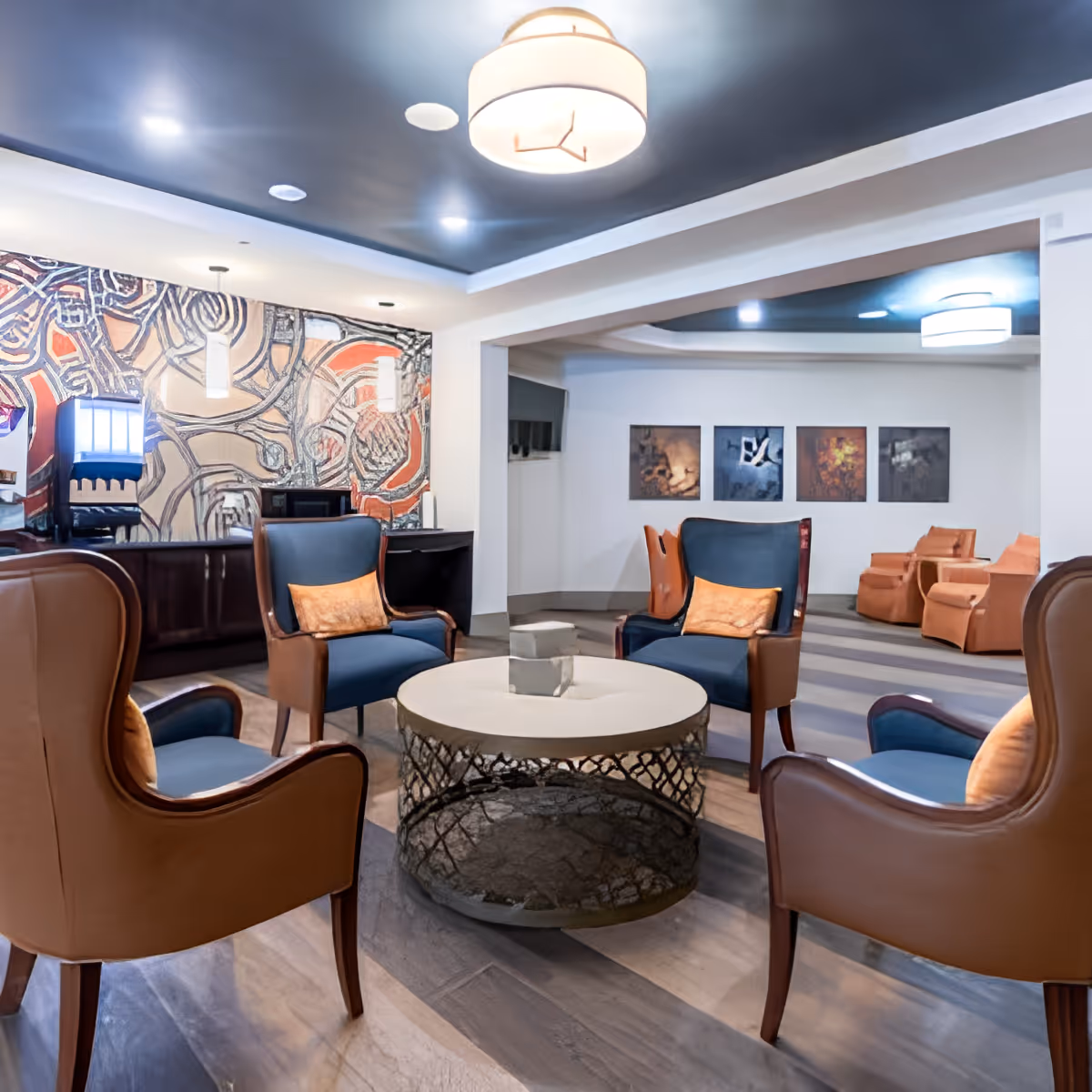 A modern lounge area with four blue and brown armchairs arranged around a circular coffee table with a decorative base. The room features a dark ceiling with recessed lighting and a large abstract mural on one wall. Additional seating with orange chairs is visible in the background along with framed artwork on the walls.
