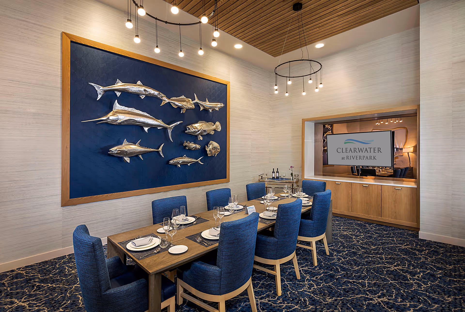 A modern dining room with a wooden table set for eight people with blue upholstered chairs. The room features a decorative wall art of silver fish on a blue background, a patterned blue carpet, and contemporary hanging light fixtures. A built-in wooden cabinet with a television screen displaying the Clearwater at Riverpark logo is visible in the background.