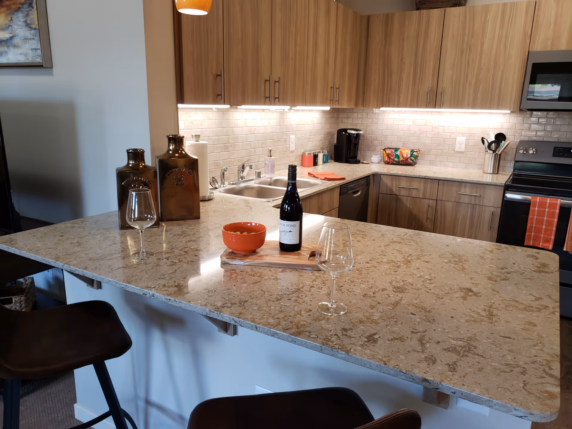 Modern kitchen with a large marble countertop island featuring two empty wine glasses, a bottle of wine, and a small orange bowl. The kitchen has wooden cabinets, a stainless steel sink, a dishwasher, a stove with an orange towel, and under-cabinet lighting. Two dark bar stools are positioned at the island.