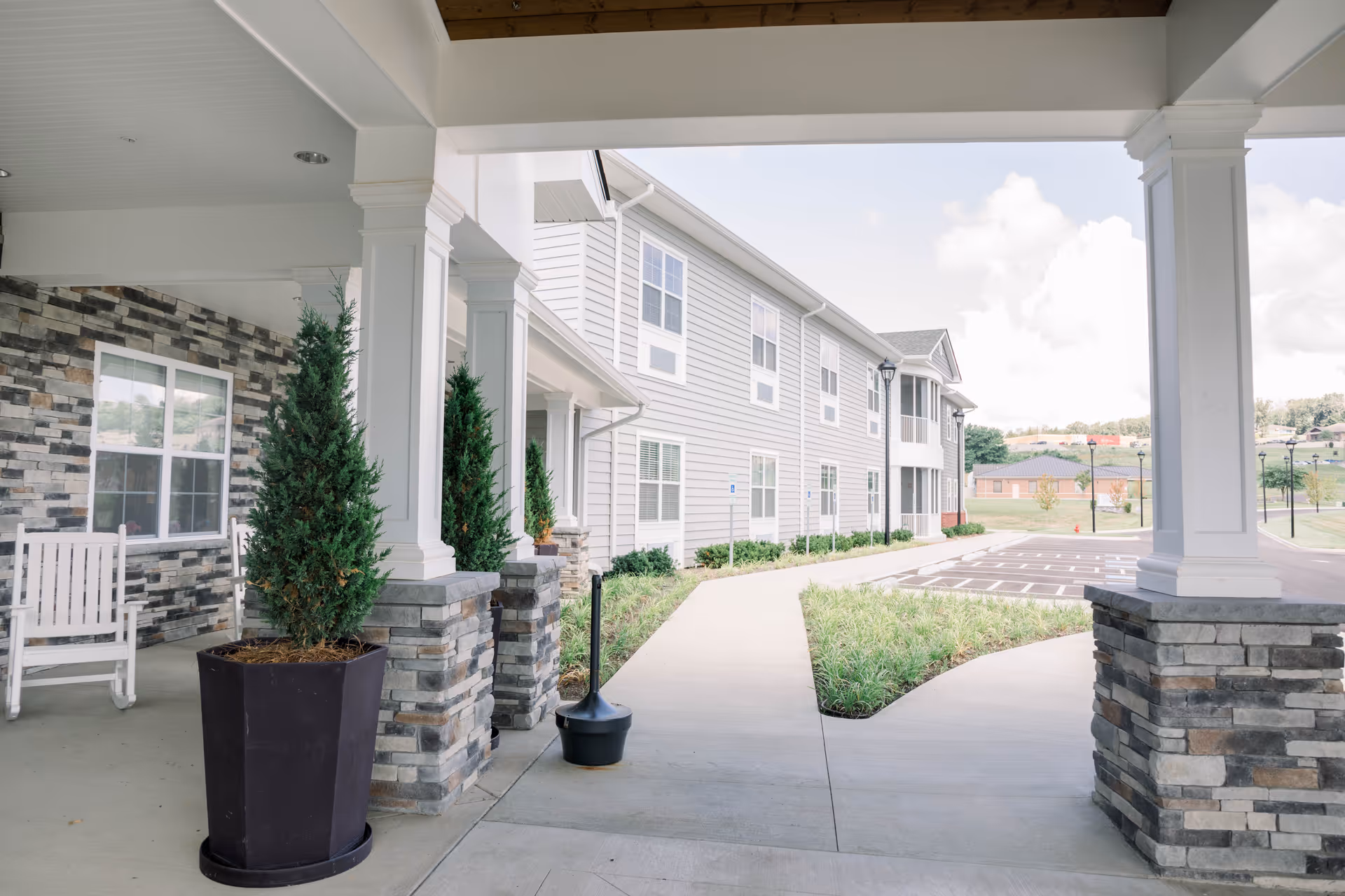 Covered front entrance and walkway of a senior living building with columns, planters, a rocking chair, and a parking area.