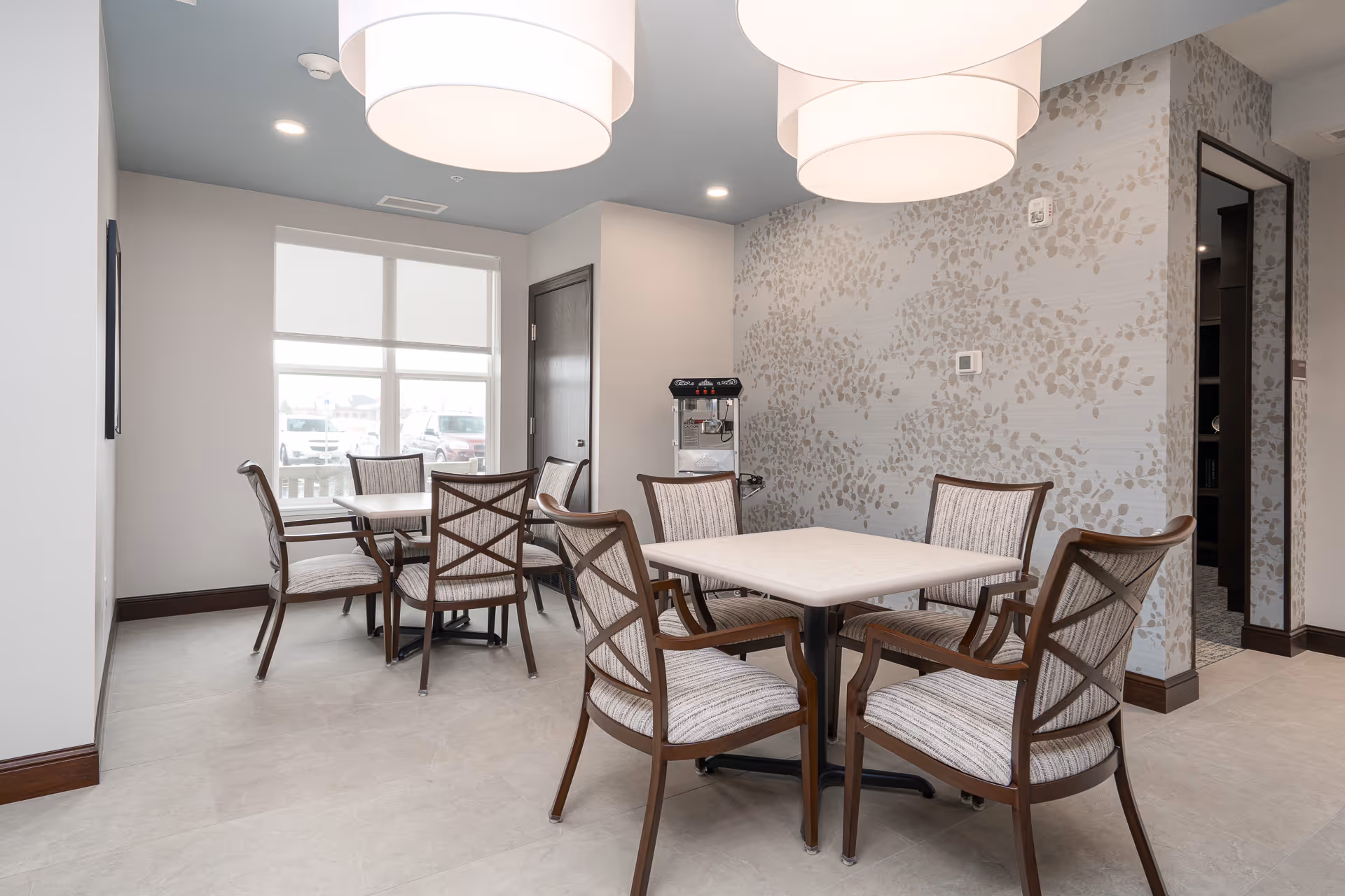 A bright dining area with two square tables, each surrounded by four cushioned wooden chairs. The room has light-colored walls, one with a subtle leaf pattern wallpaper, and a large window letting in natural light. Two large round pendant lights hang from the ceiling, and a popcorn machine is visible against the wall near a closed door.