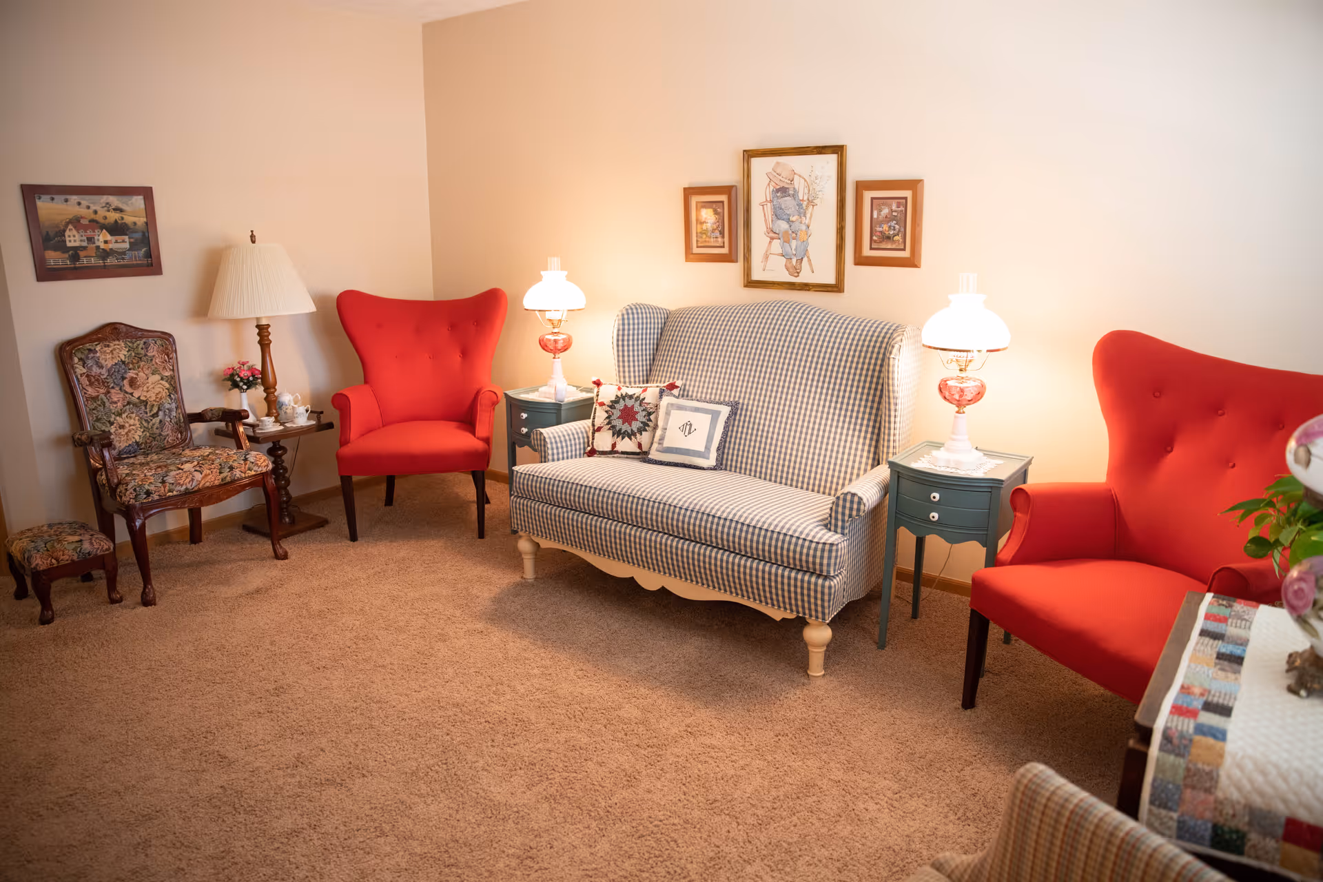 A cozy living room with beige carpet and cream-colored walls. The room features a blue and white checkered loveseat with decorative pillows, two bright red armchairs, a floral upholstered chair with a matching footstool, and two small green side tables with vintage-style lamps. There are framed pictures hanging on the wall above the loveseat.
