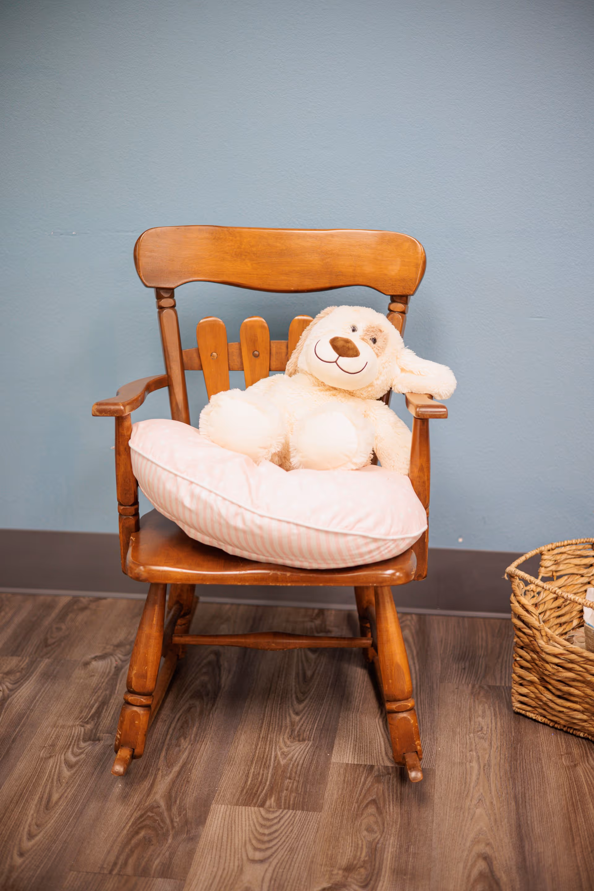 A wooden rocking chair with a pink striped pillow and a plush stuffed dog toy sitting on it, placed on a wooden floor against a light blue wall. To the right of the chair is a woven basket.