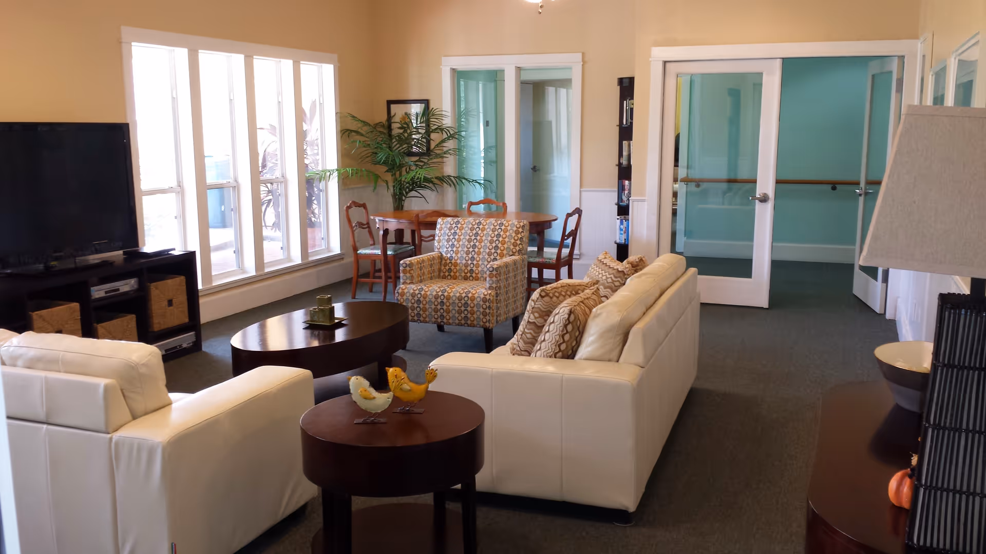 A cozy living room area with white leather sofas, a patterned armchair, and two round wooden coffee tables. There is a large flat-screen TV on a black stand with storage baskets underneath. A round wooden dining table with chairs is placed near large windows letting in natural light. The walls are painted a warm beige color, and there are glass double doors leading to another room. A potted plant and a table lamp add decorative touches.
