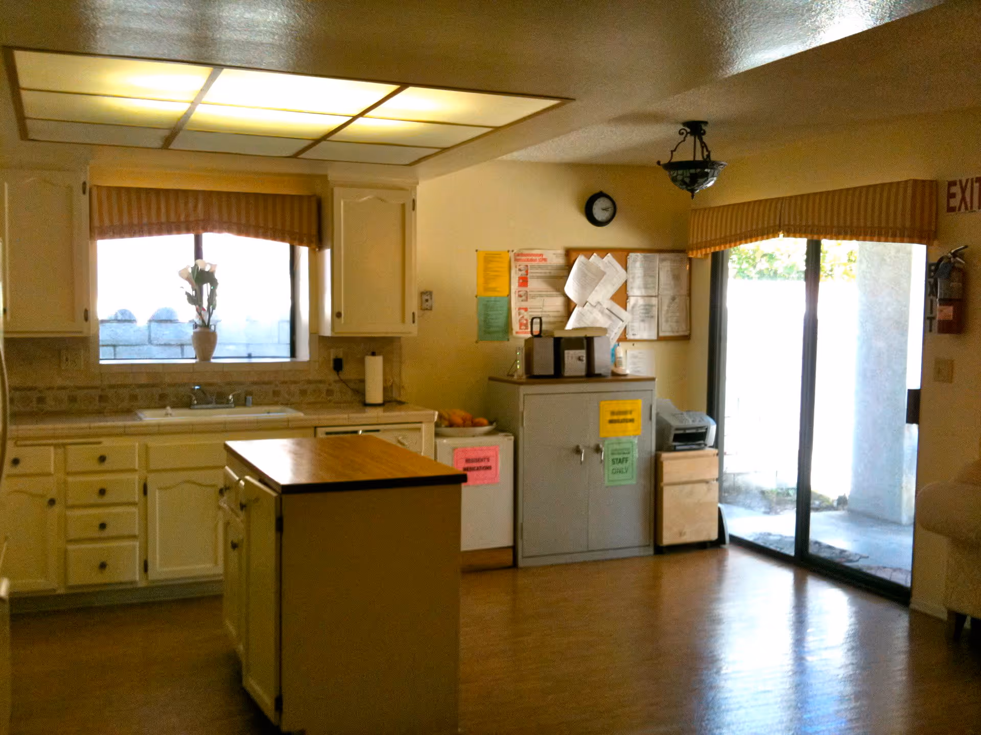 Interior view of a kitchen area with white cabinets, a central island with a wooden countertop, a window with a flower vase on the sill, and a sliding glass door leading outside. There are various notices and papers pinned on a bulletin board above a cabinet, and an exit sign is visible near the door.