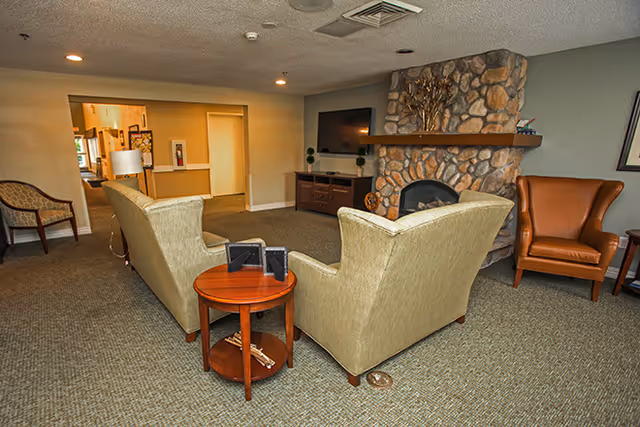 A cozy living room area in a senior living facility featuring two beige armchairs facing a stone fireplace with a mounted flat-screen TV above a wooden cabinet. There is a small round wooden side table between the chairs, and a brown leather armchair is positioned to the right. The room has carpeted flooring and neutral-colored walls with recessed lighting in the ceiling.