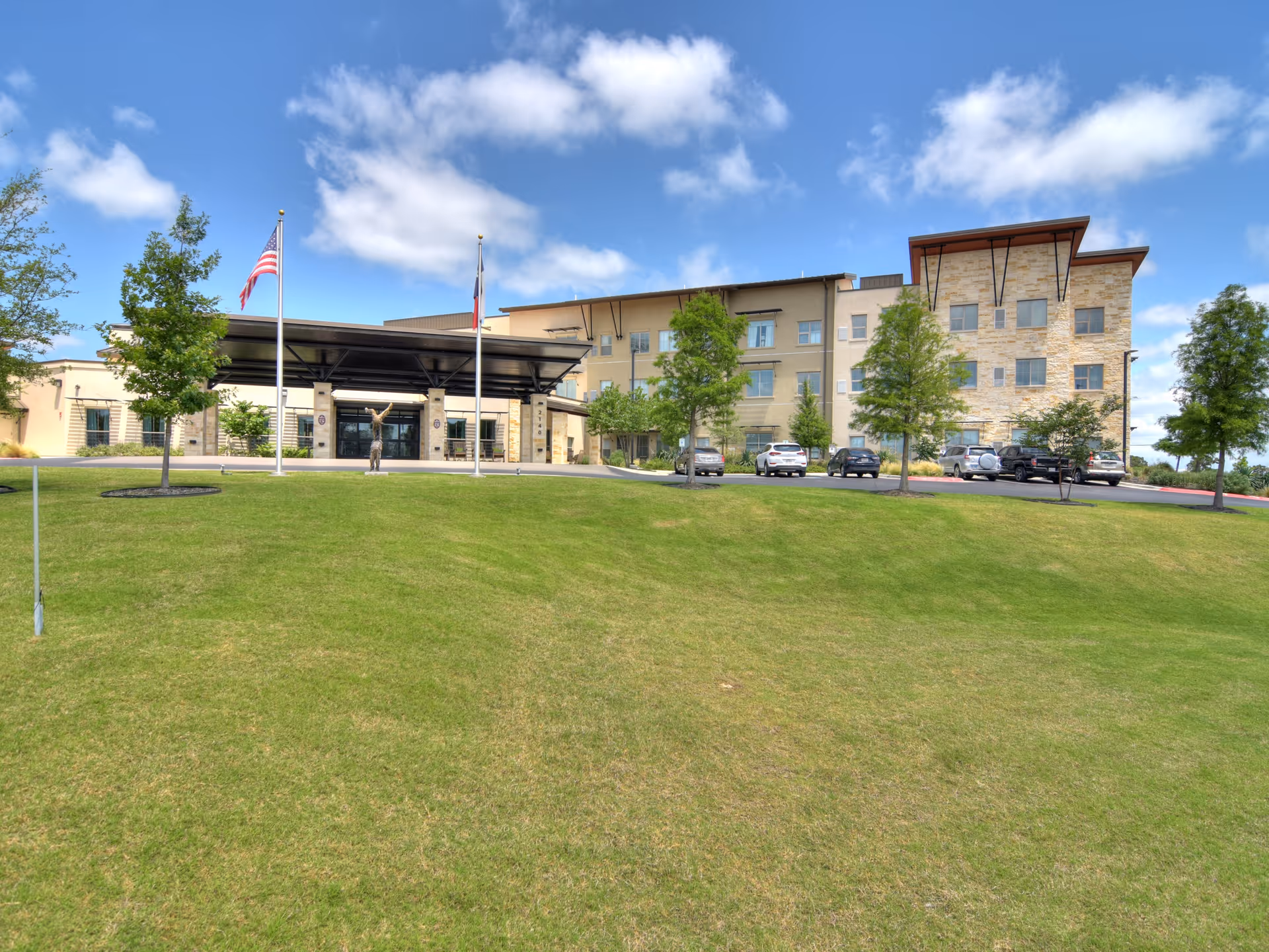 Exterior view of Élan Westpointe at New Braunfels senior living facility showing a modern multi-story building with stone and beige walls, a covered entrance with two flagpoles flying the American and Texas flags, several trees, parked cars, and a well-maintained grassy lawn under a partly cloudy blue sky.