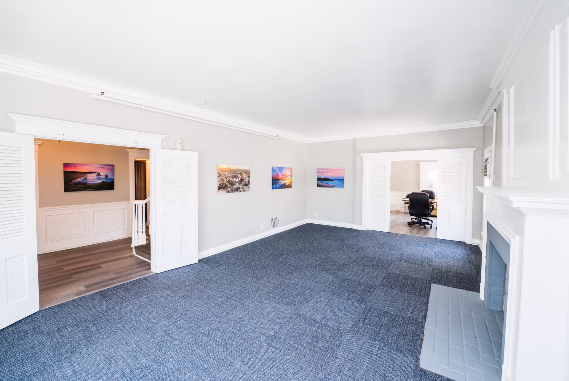 A spacious, empty room with blue carpet and light gray walls featuring four colorful landscape paintings. The room has white trim and a white fireplace on the right side. Two doorways with white double doors lead to adjacent rooms, one showing a staircase and the other an office area with black chairs and a desk.