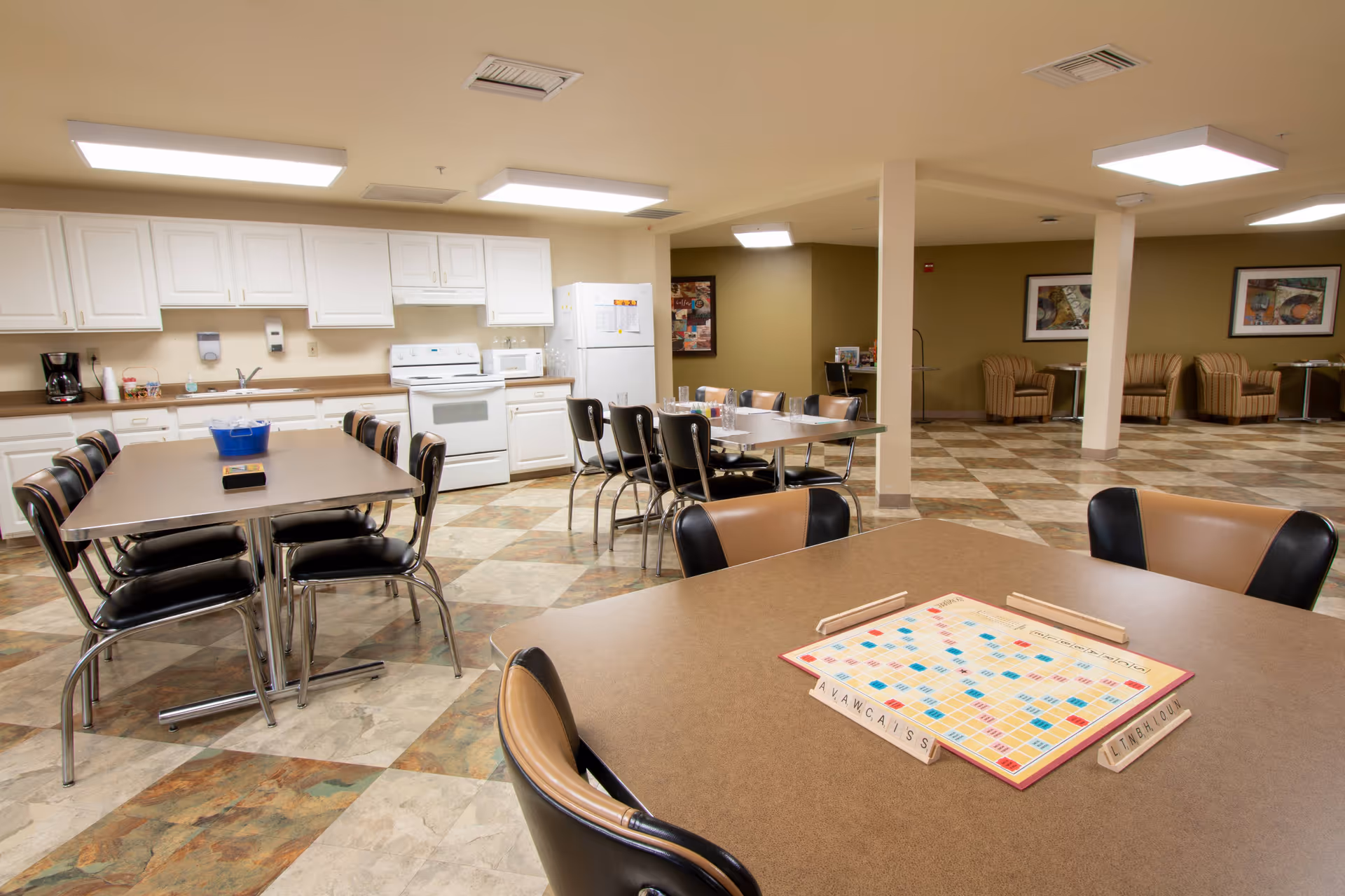 A spacious common area in a senior living facility featuring a kitchen with white cabinets, a stove, microwave, refrigerator, and coffee maker. Several tables and chairs are arranged for dining or activities, with one table in the foreground set up with a Scrabble board game. The room has tiled flooring and neutral-colored walls with framed artwork and comfortable seating areas in the background.