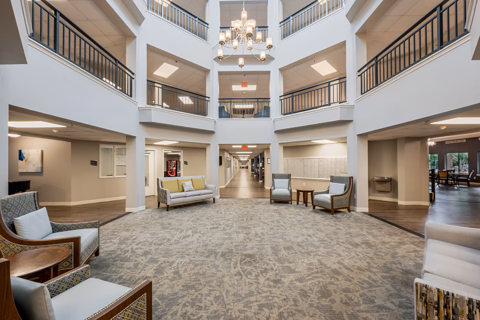A spacious, well-lit common area in a senior living facility with multiple seating arrangements including armchairs and sofas. The area features a high ceiling with a chandelier, railings on the upper floors, and a long hallway leading to other rooms. There is a water fountain and a wall of mailboxes visible on the right side.