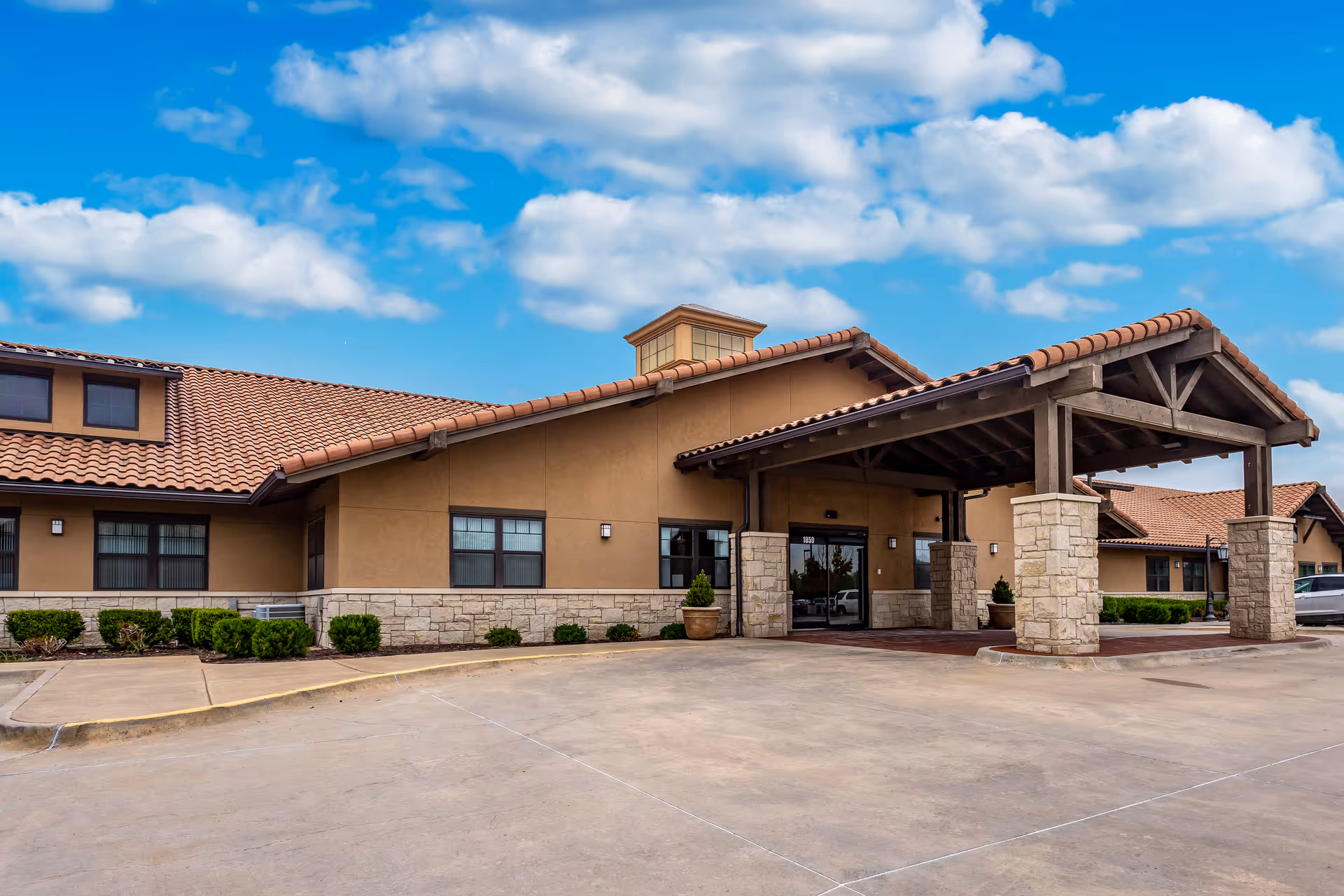 Exterior view of Chisholm Place facility showing a single-story building with a tiled roof, stone and stucco walls, a covered entrance supported by stone pillars, and a clear blue sky with scattered clouds.