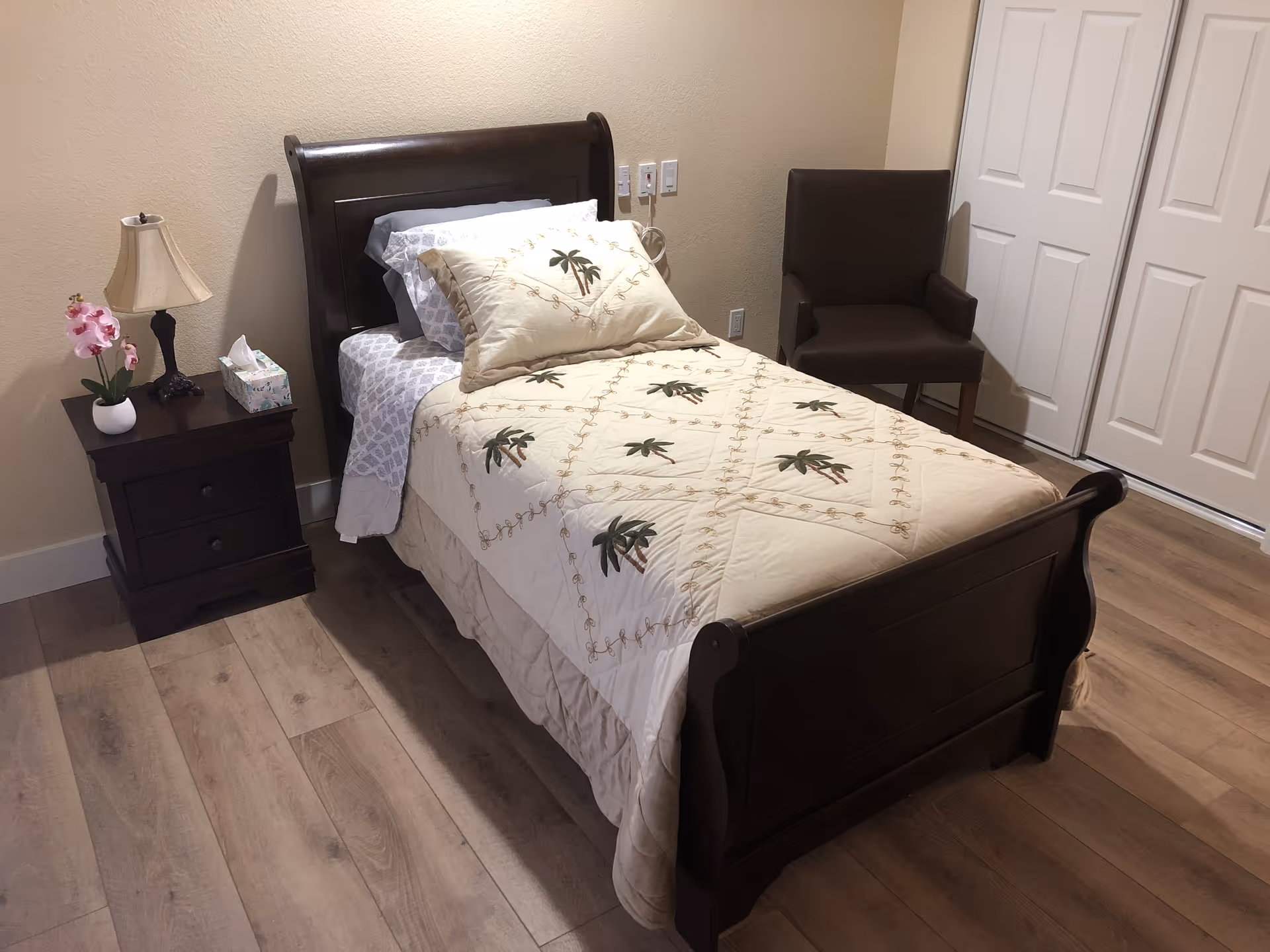 A tidy single bedroom with a wooden sleigh bed made with a patterned quilt, a bedside table with lamp and flowers, a chair, and a closet.