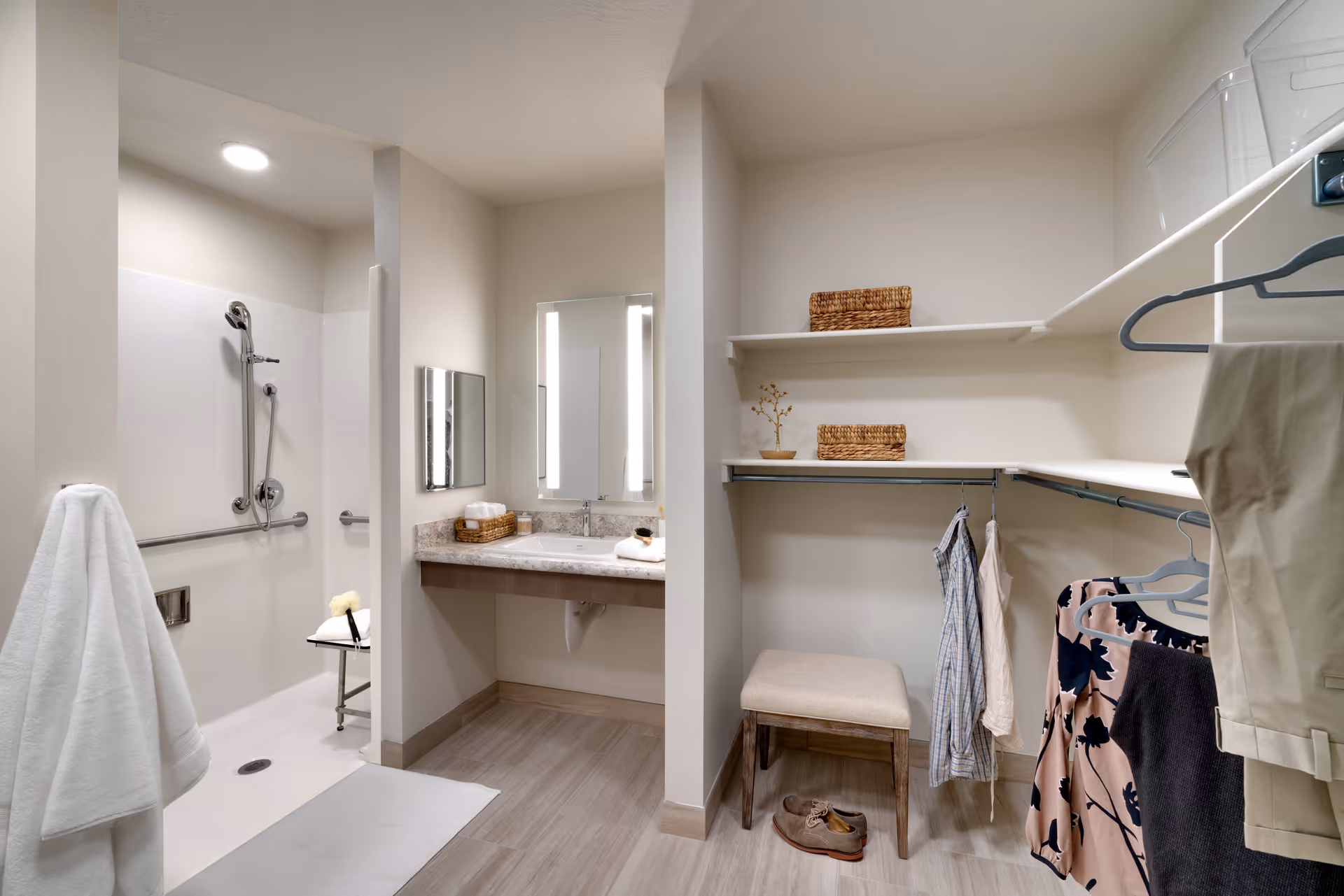 A senior living facility bathroom and dressing area featuring a walk-in shower with grab bars and a shower chair, a vanity with a sink and illuminated mirror, and an adjacent open closet space with hanging clothes, shelves with baskets, a small cushioned bench, and a pair of shoes on the floor.