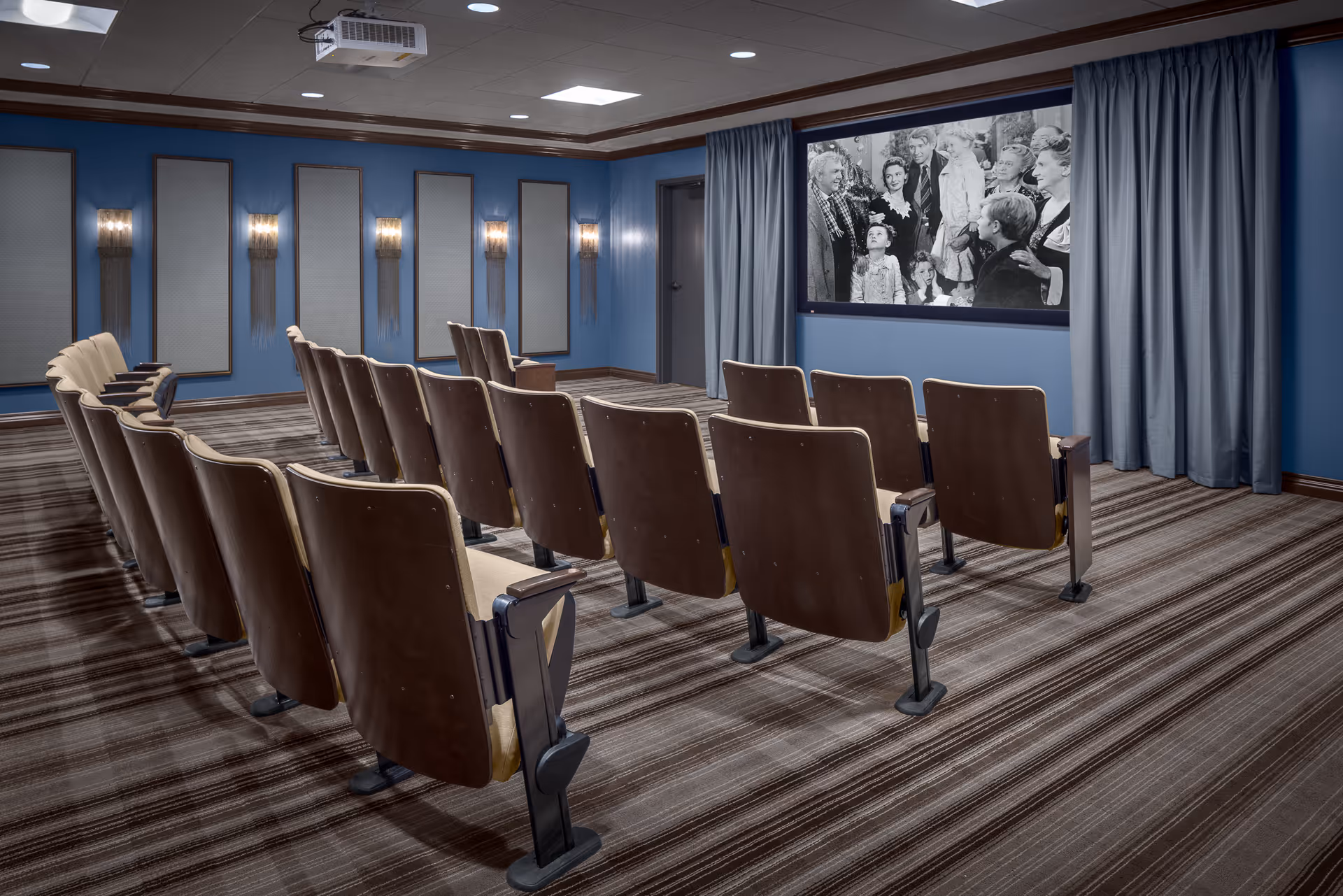 A small theater room with three rows of brown cushioned seats facing a large screen showing a black and white movie. The walls are painted blue with decorative panels and wall sconces, and the floor is covered with striped carpet. A projector is mounted on the ceiling.