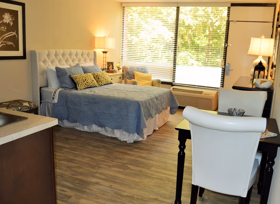 A bright studio bedroom with a bed, side tables and lamps, a windowed seating area, and a small dining desk and kitchenette.